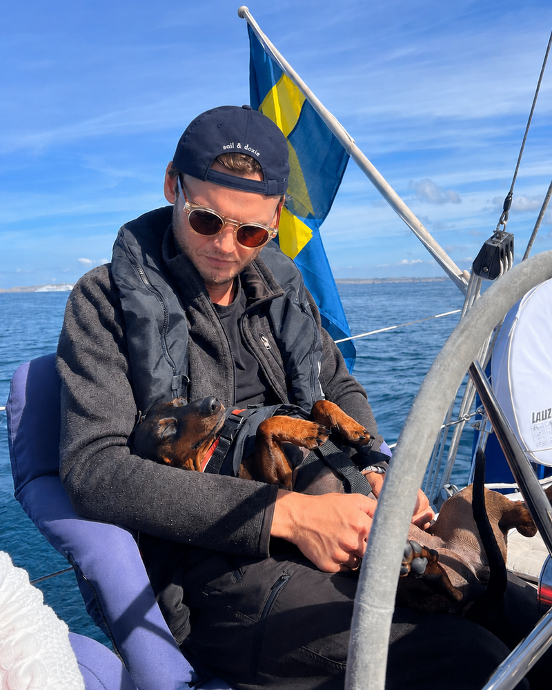 A man wearing sunglasses, a black cap, and a dark fleece jacket sits on a boat holding a small brown dog. A blue and yellow Swedish flag is visible in the background under a blue sky.