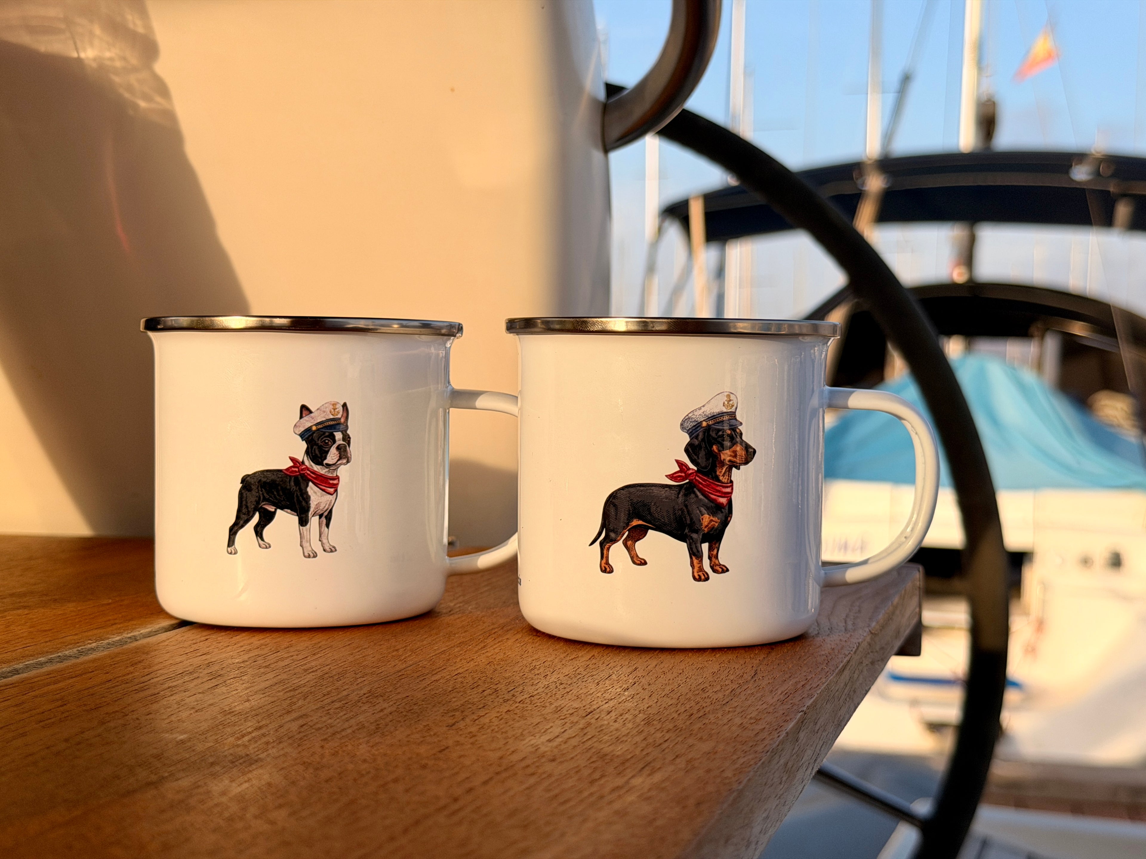 Two white enamel camping mugs with silver rims sitting on a wooden table outdoors, featuring illustrations of a black and white dog wearing a top hat and a dachshund wearing a top hat and red bandana. A sailboat is blurred in the background.