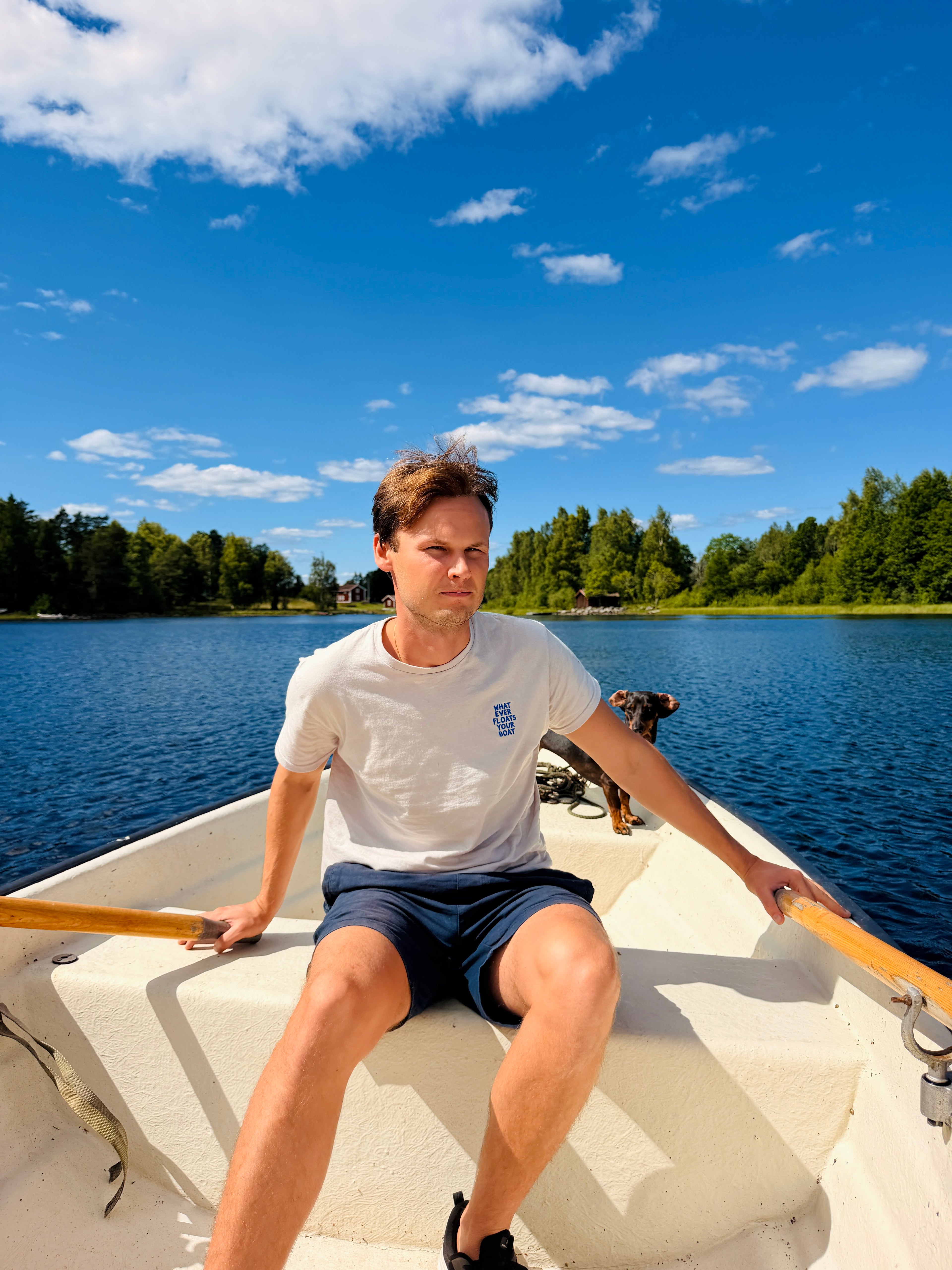 Man rowing a boat on a calm lake with trees in the background. A small dog is sitting in the boat behind the man.