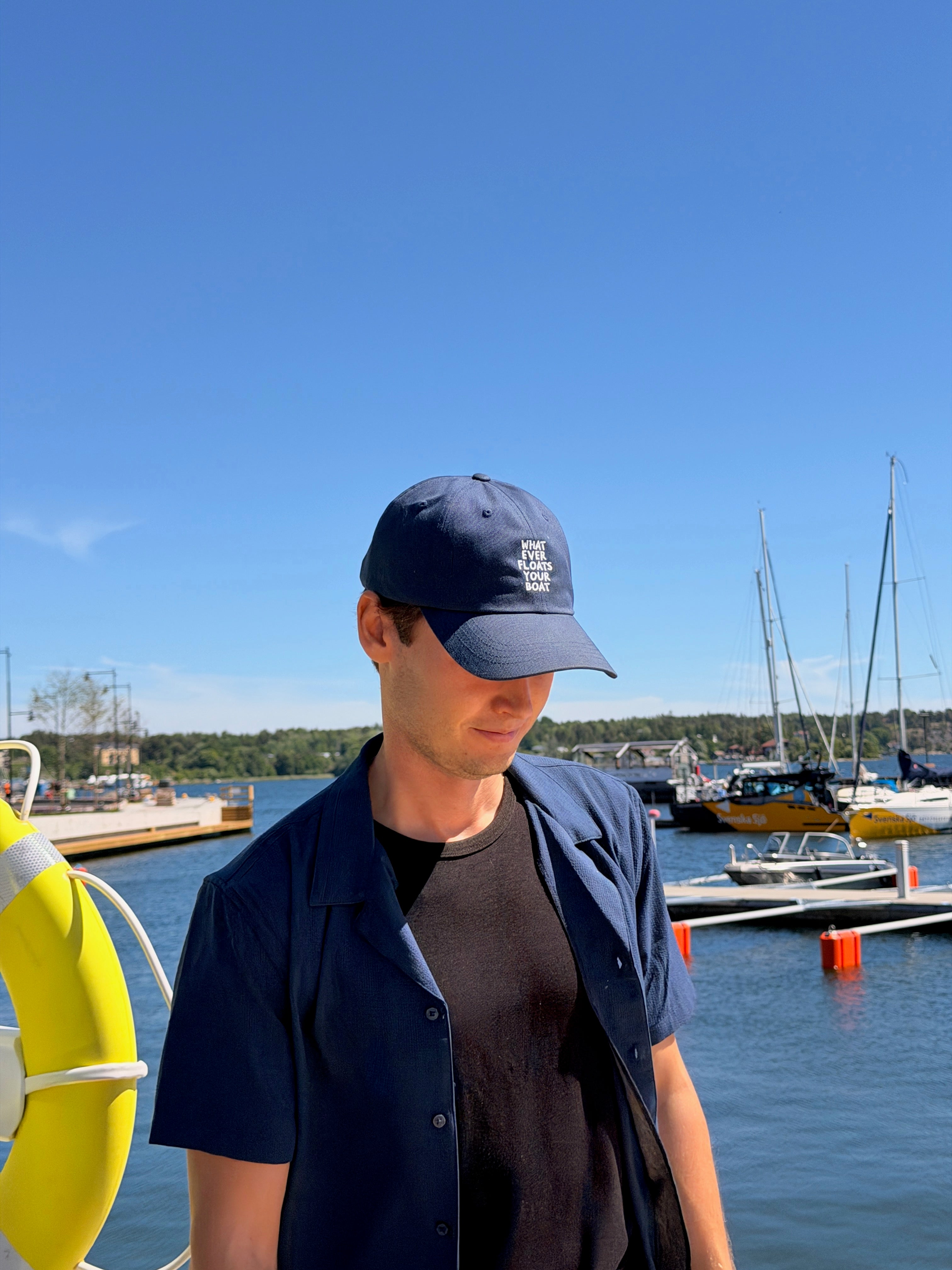 A man wearing a navy blue baseball cap with a white embroidered logo, a navy short-sleeve button-up shirt, and a black t-shirt, standing on a dock with a marina and blue sky in the background.