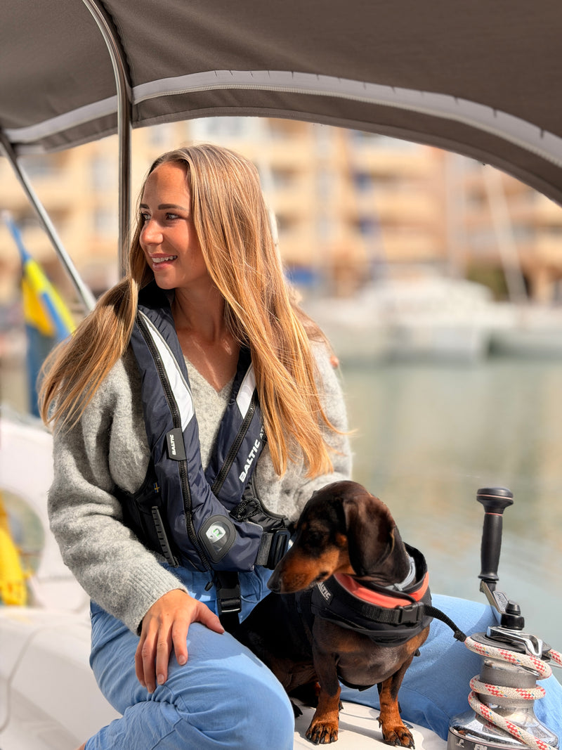 A woman on a boat wearing a black and navy life jacket, sitting next to a small dachshund wearing a life vest, with a marina background.