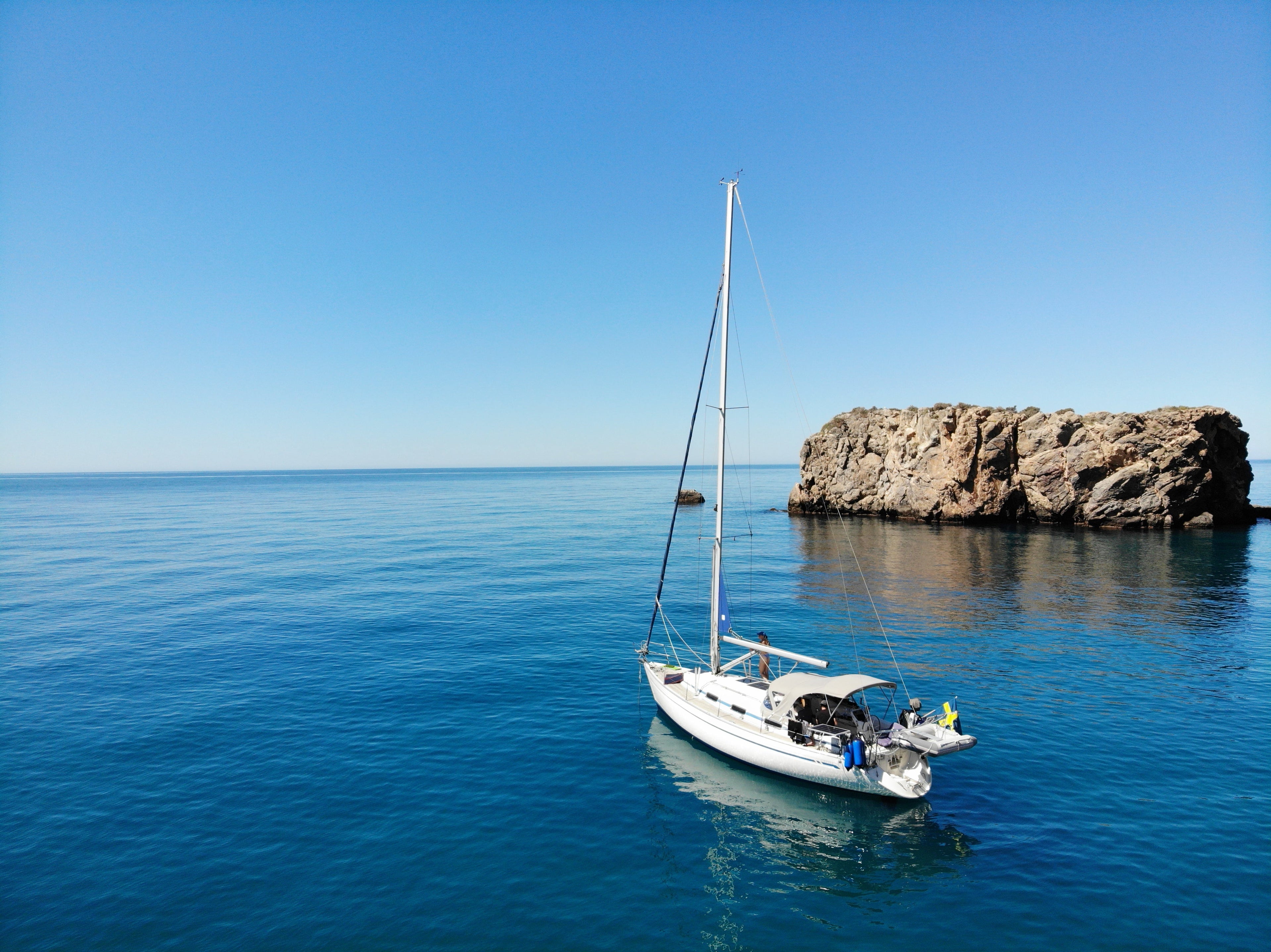A white sailboat with its mast raised is anchored in calm blue water next to a rocky outcrop under a clear blue sky.
